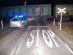 Na zdjęciu radiowóz z włączonymi sygnałami uprzywilejowania, w tle pociąg i znaki drogowe