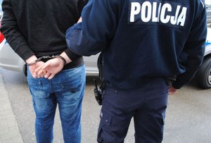 Fotografia kolorowa przedstawiająca umundurowanego policjanta który prowadzi zatrzymanego mężczyznę. Mężczyzna ma założone kajdanki na ręce trzymane z tyłu.