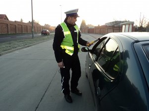 na fotografii policjant przy kontrolowanym pojeździe podaje kierowcy urządzenie do badania na zawartość alkoholu w wydychanym powietrzu