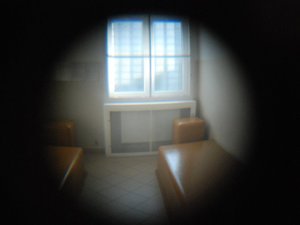 Kolorowa fotografia. Widok przez wziernik celi w policyjnych aresztach. Dwie prycze ze stołami i okno z kratami.