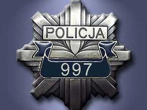 Kolorowa fotografia. Policyjna odznaka na niebieskim tle z napisem Policja i numerem 997.