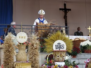 Kolorowa fotografia. Biskup Andrzej Jeż wygłasza homilię z ambony ołtarza polowego. Przed ołtarzem wieńce dożynkowe ustawione w rzędach.