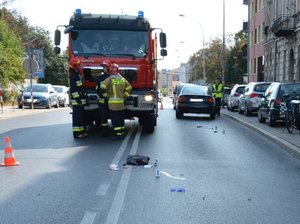 Fotografia przedstawia miejsce wypadku drogowego w Jarosławiu na ul. 3-go Maja. Na zdjęciu znajduje się na środku jezdni wóz strażacki. W tle po lewej stronie na drodze stoi samochód osobowy,