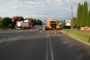 na fotografii w oddali widać samochód ciężarowy koloru pomarańczowego, oraz stojącego obok opla, w tle widać także wóz strażacki i strażaków, a także pachołki wyznaczające zamknięcie drogi