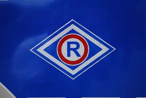 Oznaczenie służby ruchu drogowego, duża litera R wpisana w niebiesko-biały romb umieszczony na granatowym tle.