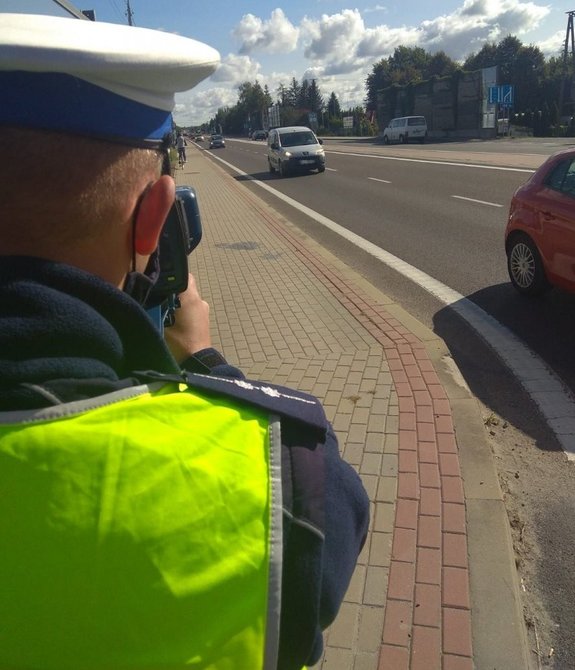 Policjant, który w ręku trzyma urządzenie do mierzenia predkości. Stoi przy ulicy, w tle jadące pojazdy