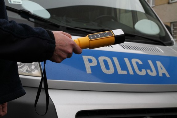 policjant trzymający w ręku urządzenie do badania stanu trzeźwości. w tle radiowóz z napisem na masce policja