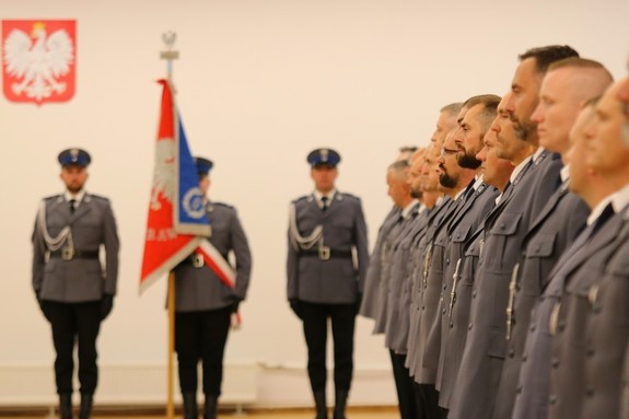 Stojący w rzędzie awansowani policjanci, na drugim planie poczet sztandarowy w czasie uroczystości