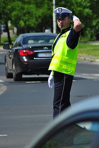 Policjant ruchu drogowego, kierujący ruchem, w tle jadący samochód. Pora dzienna.