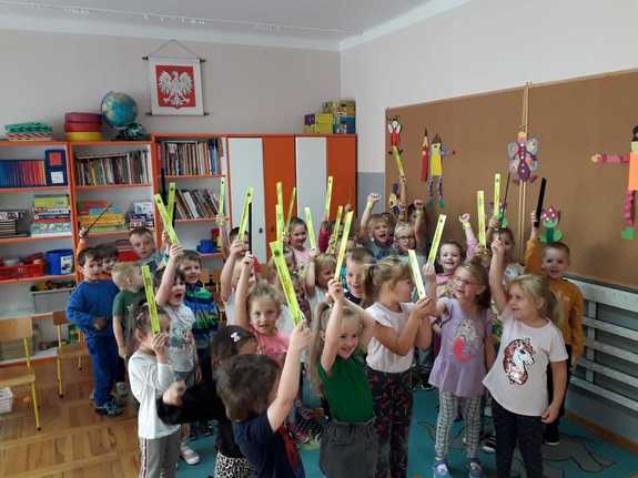 Zdjęcie kolorowe przedstawia dzieci w sali przedszkolnej , które w raczkach wyciągniętych ku górze trzymaj elementy odblaskowe