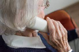 Zdjęcie kolorowe przedstawia starsza kobietę która w ręku przy uchu trzyma słuchawkę od aparatu telefonicznego stacjonarnego