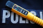 Zdjęcie kolorowe przedstawia urządzenie do pomiaru alkoholu w wydychanym powietrzu „Alcoblov” w kolorze żółtym z czarnym ustnikiem. Urządzenie jest położone na granatowym tle z napisem białym „POLICJA”