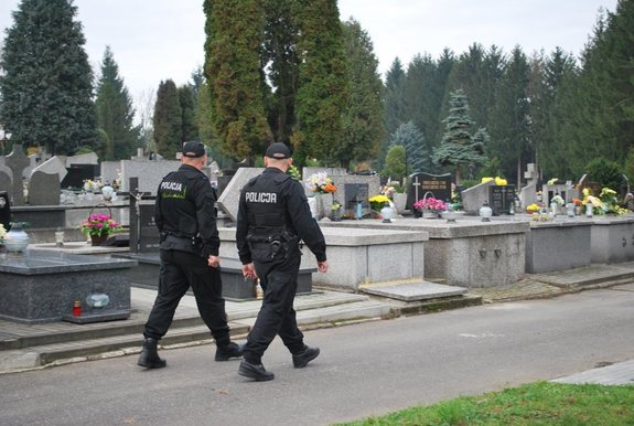 Dwóch policjantów w czarnych mundurach podczas patrolu terenu cmentarza. W tle widok nekropolii