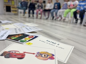 Na zdjęciu na podłodze leza kolorowe kartki z ilustracjami służące do nauki zasad bezpiecznego poruszania się po drogach. W tel widać siedzących na białych krzesełkach przedszkolaków.