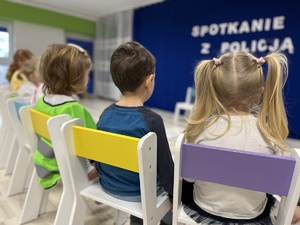 Na zdjęciu w sali tyłem na białych krzesełkach z kolorowymi oparciami siedzą dzieci. Przed nimi na granatowej scianie widać napis: SPOTKANIE Z POLICJĄ.