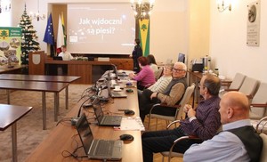 Na pierwszym planie seniorzy siedzący przed ekranami laptopów, w tle policjantka wyświetlająca prezentację multimedialną dotyczącą bezpieczeństwa w ruchu drogowym. Obok niej unijne flagi, a także gminy Jedlicze i Polski.
