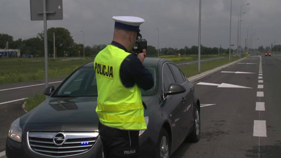 zdjęcie przedstawia umundurowanego policjanta stojącego w rejonie drogi i mierzącego prędkość nadjeżdżającego pojazdu