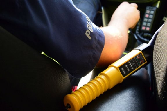 na zdjęciu widoczne jest urządzenie do badania stanu trzeźwości w kolorze żółtym- tuba. urządzenie umieszczone jest w radiowozie. widoczny fragment wnętrza auta i umundurowanego funkcjonariusza Policji