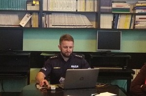 Umundurowany policjant przed ekranem komputera podczas prowadzonego spotkania. W tle półki ścienne z segregatorami i książkami