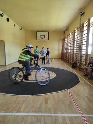 Na sali gimnastycznej chłopiec w kasku jedzie na rowerze. W oddali widać uczniów, którzy stoja.