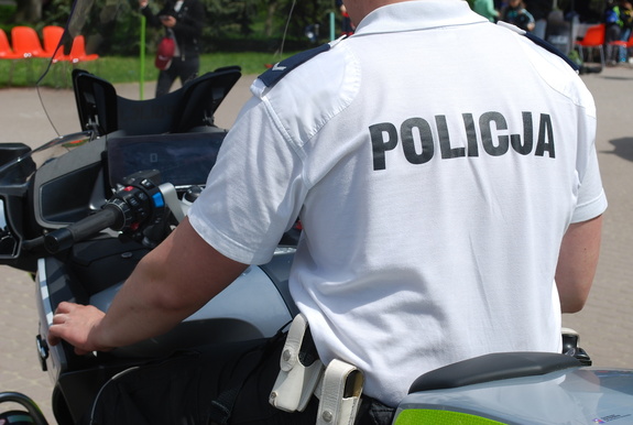 Na zdjęciu siedzący tyłem na motocyklu służbowym  policjant ruchu drogowego w białej koszulce polo z napisem policja na plecach.