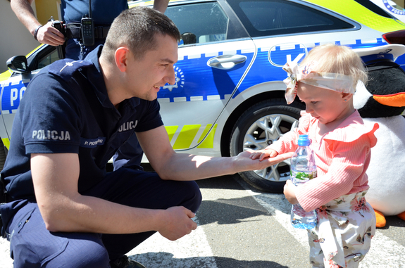 Policjant podający rękę małej dziewczynce. W tle oznakowany radiowóz.