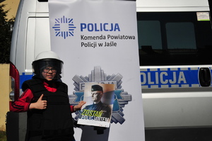 Chłopiec ubrany w sprzęt do zabezpieczeń zgromadzeń na tle banneru oraz z plakatem z napisem zostań policjantem