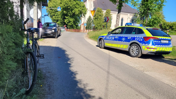 Miejsce wypadku drogowego w miejscowości Nowosielce. Na zdjęci po lewej stronie radiowóz oznakowany. Po prawej widoczny jest samochód osobowy czarny. Tuż przed nim rower. W tle kościół oraz drzewa.