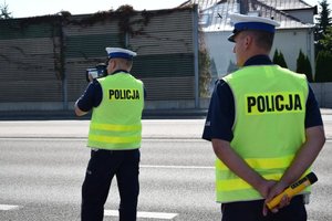 Dwaj policjanci ruchu drogowego w kamizelkach odblaskowych z napisem POLICJA. Policjant stojący po lewej stronie trzyma w rękach miernik prędkości.
