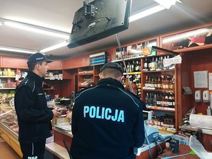 Dwaj policjanci stoją przy ladzie sklepu monopolowego. Sprawdzają dokumenty.