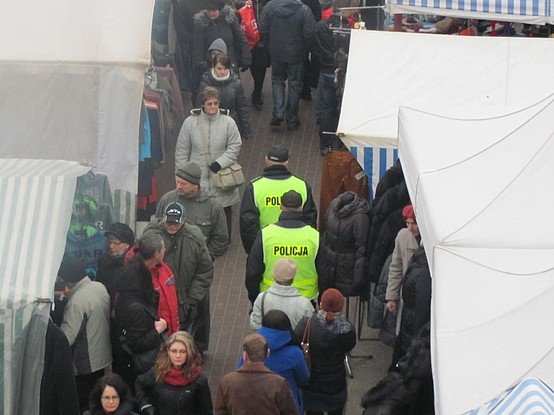 Targowisko. Ludzie spacerujący między straganami. W środku patrolujący teren bazaru policjanci.