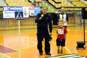 Umundurowany policjant trzyma mikrofon obok stoi chłopiec w wieku 6 - lat, ubrany w czerwoną koszulkę z krótkim rękawem i ciemne spodenki.