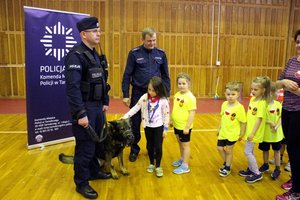 Dwaj umundurowani policjanci z psem policyjnym. W tle baner z napisem Policja. Obok stoją przedszkolaki ubrane w żółte koszulki.