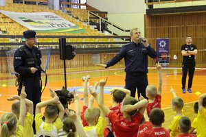 Grupa przedszkolaków siedzących na podłodze w hali sportowej. W tle dwaj  umundurowani policjanci.