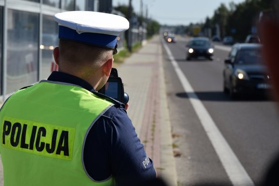 Na zdjęciu policjant ruchu drogowego trzymający urządzenie do pomiaru prędkości, w tle droga i jadące samochody.