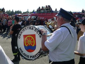 Na pierwszym planie jeden z członków Orkiestry Dętej z Miejsca Piastowego w tle strażacy oraz wozy strażackie.