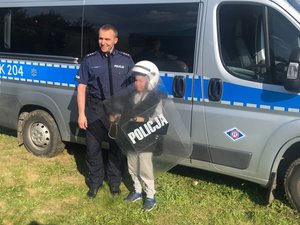 Policjant st. asp. Paweł Buczyński wspólnie z chłopcem ubranym w  elementy umundurowania wykorzystywanego do zabezpieczania imprez masowych na tle radiowozu strażackiego.
