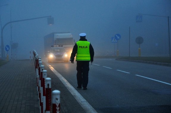 Na zdjęciu policjant idący drogą. W tle widać radiowóz i samochód ciężarowy. Panują trudne warunki atmosferyczne: mgła i opady deszczu.