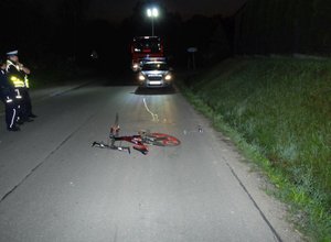 Fotografia kolorowa wykonana nocną porą. Na zdjęciu widoczna jest jezdnia. Na środku drogi znajduje się rower w kolorze czerwonym. Po lewej stronie zdjęcia znajdują się dwaj policjanci. W tle zdjęcia widoczny jest radiowóz a za nim pojazd straży pożarnej.
