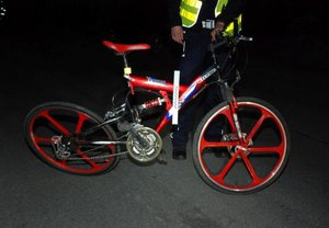 Fotografia kolorowa wykonana nocną porą. Na zdjęciu widoczny jest policjant ubrany w kamizelkę odblaskową. Funkcjonariusz trzyma rower koloru czerwonego do którego przykłada skalówkę.
