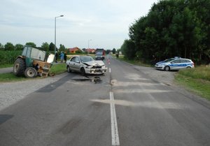 Zdjęcie przedstawia miejsce kolizji drogowej. Po lewej stronie w rowie znajduje się uszkodzony ciągnik. Na jezdni stoi uszkodzona mazda. Po prawej stronie jezdni widoczny jest radiowóz.