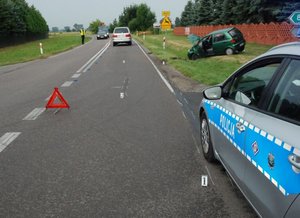 Na zdjęciu widać miejsce wypadku drogowego. Po prawej stronie znajduje się radiowóz. Za radiowozem na trawiastym poboczu widoczny jest uszkodzony samochód koloru zielonego. Na środku drogi znajduje się trójkąt ostrzegawczy.