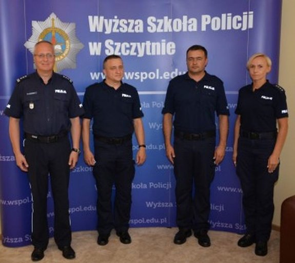 Fotografia przedstawia czterech umundurowanych policjantów. W tle widnieje napis o treści Wyższa Szkoła Policji w Szczytnie.