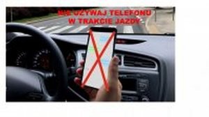 Zdjęcie przedstawia kierującego pojazdem który w ręce trzyma telefon komórkowy. Telefon ten jest przekreślony. Nad telefonem widnieje napis w kolorze czerwonym o treści &quot;Nie używaj telefonu w trakcie jazdy&quot;