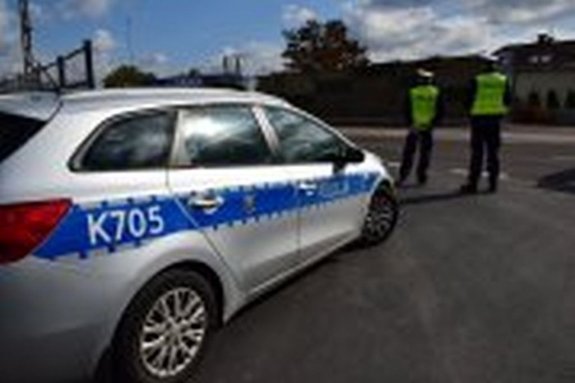 Fotografia przedstawia radiowóz. Przed pojazdem widoczni są dwaj policjanci ruchu drogowego, którzy dokonują pomiaru prędkości kierujących.