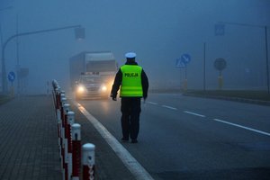 Na zdjęciu umundurowany policjant dbający o bezpieczeństwo na drodze, w tle policyjny radiowóz oraz samochód ciężarowy. Pogoda jest pochmurna, panuje gęsta mgła.