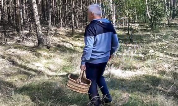 Starszy mężczyzna idący z wiklinowym koszykiem na grzyby. W tle las liściasty.