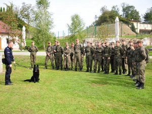 na zdjęciu stojący w dwuszeregu żołnierze WOT  a przed nimi policjant - przewodnik psa służbowego z psem o imieniu Herc