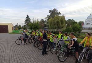 Zdjęcie kolorowe . Umundurowana policjanta ruchu drogowego przekazuje dzieciom ze Szkoły Podstawowej w Piskorowicach opaski odblaskowe. Dzieci  stoją z rowerami na palcu szkolnym.
