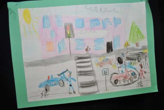 Plakat- rysunek przedstawia przejście dla pieszych przed budynkiem szkoły, w tle widać narysowany radiowóz oraz inne pojazdy wraz z uczestnikami ruchu.